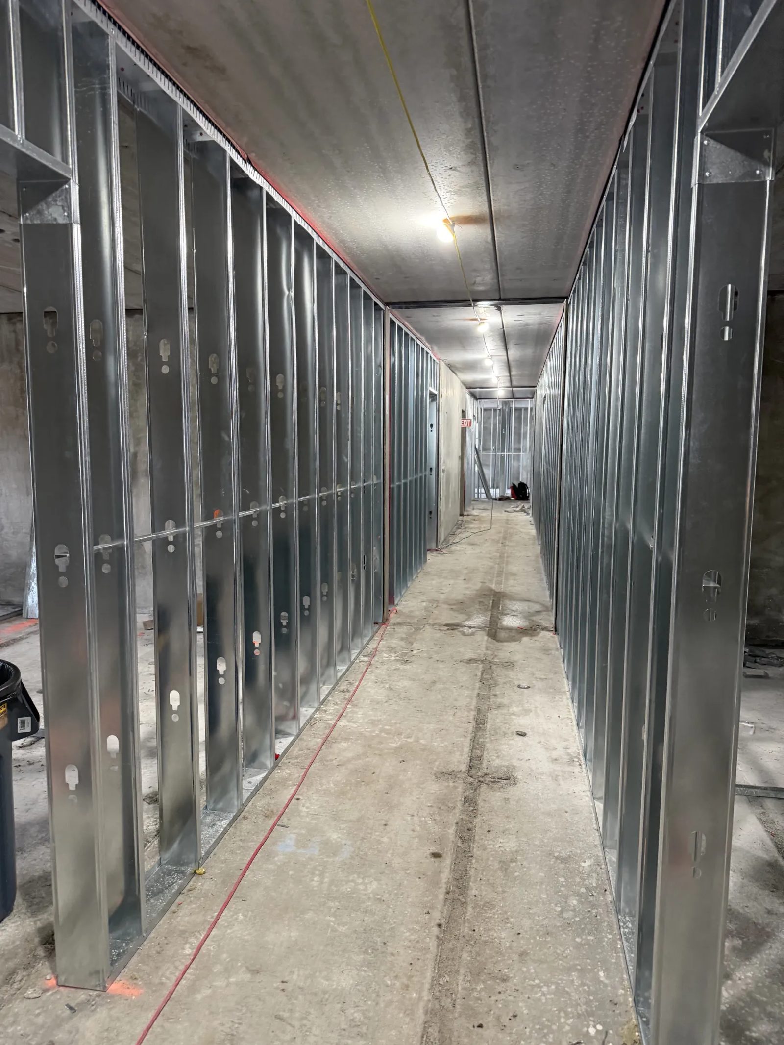 Long interior corridor under construction with metal stud framing along both sides and an exit sign visible ahead