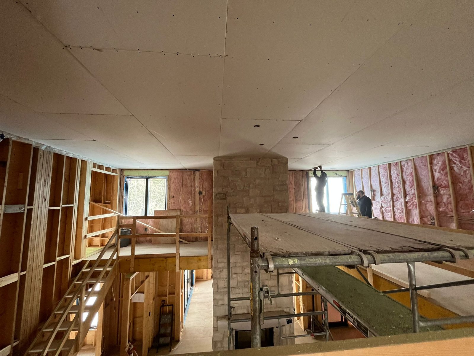 Two crew members on scaffolding taping and finishing the vaulted ceiling of a large custom home, with framed walls and a stone chimney visible below